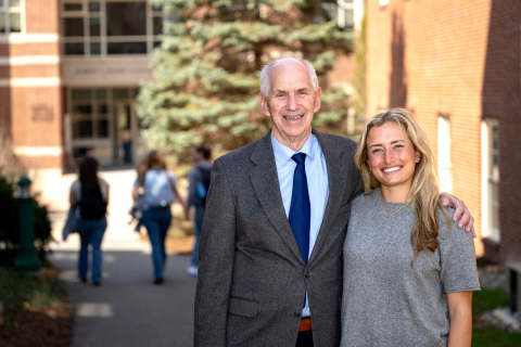 Nicole Lioumis wears a gray short-sleeve shirt and smiles on campus while standing next to Edward McDevitt '72.
