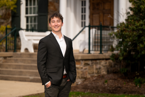 Matthew Karic wears a black suit and white shirt and smiles while standing in front of a building with steps and a white bench.