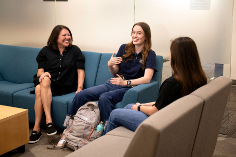 Maya Fetzer sits on a blue couch and talks with two other people while smiling at the camera.