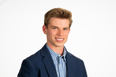 Tyler Thrutchley '26 wears a light blue shirt with a dark blue blazer and smiles against a white backdrop