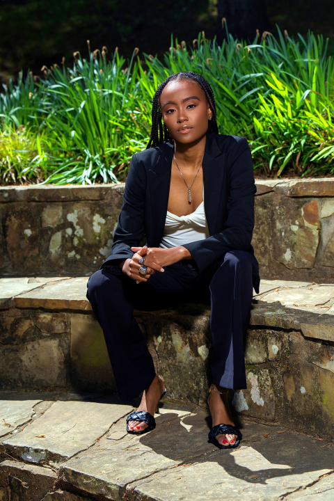 Gia Hayes sits on a stone step while wearing a navy pants suit, with green tall grass in the background