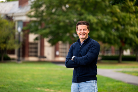 Freddie Lapworth wears a blue sweater and jeans and smiles with arms crossed on campus
