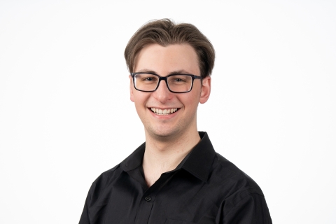 Joe Dox wears black rimmed glasses and a black collard shirt and smiles against a white backdrop.