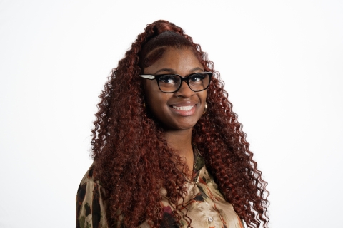 Tahadai Crews-Harris smiles wearing black-rimmed glasses and a brown top, against a white backdrop.