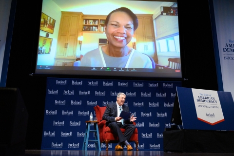 Condoleezza Rice appears on a video screen above president John Bravman, sitting on a chair on stage.