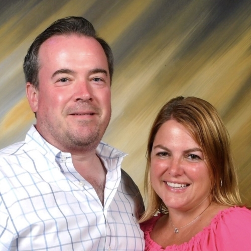 Portrait of a smiling couple. A man is on the left in a white and black plaid button down. A woman is on the right in a pink top.